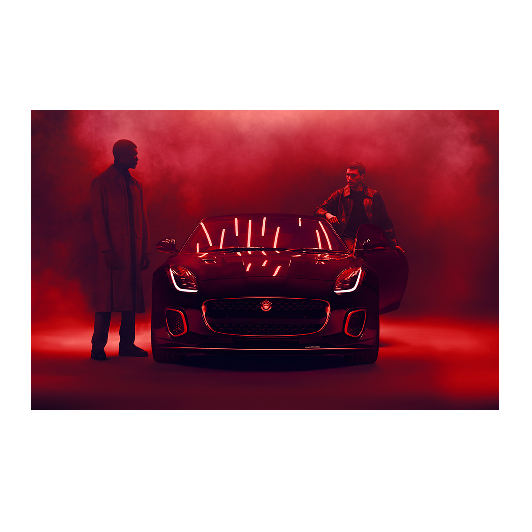 A dark-colored Jaguar sports car is showcased with two men in a red-lit environment, highlighting automotive design and style.