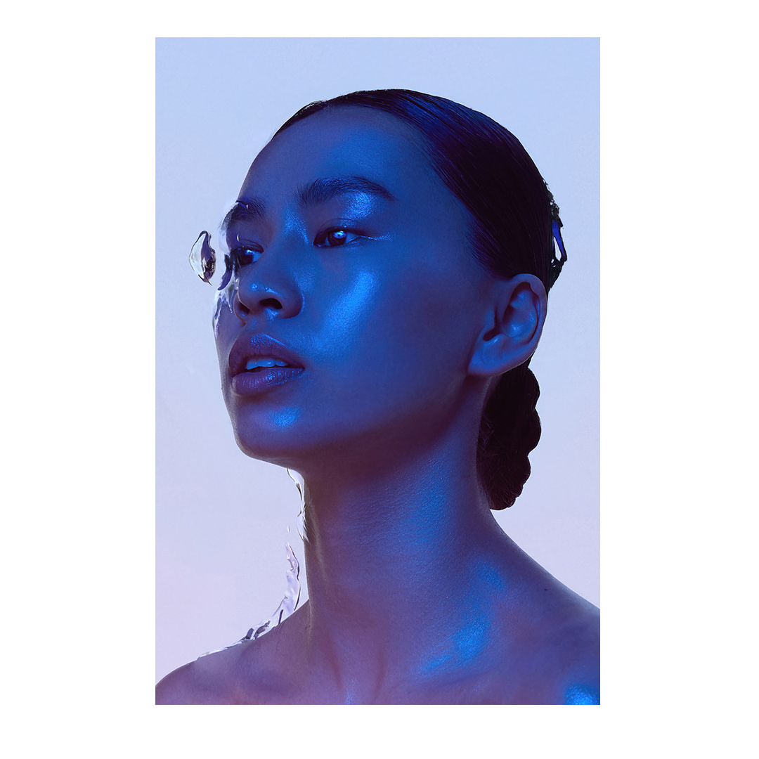 Portrait of Asian woman with glowing skin and water droplets, captured in beauty shot with blue lighting and a light background.