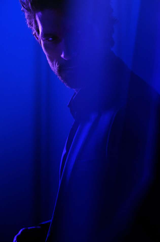 Dramatic portrait of a bearded adult male in a dark blue suit, partially obscured by a blue light leak effect. Moody indoor portrait.