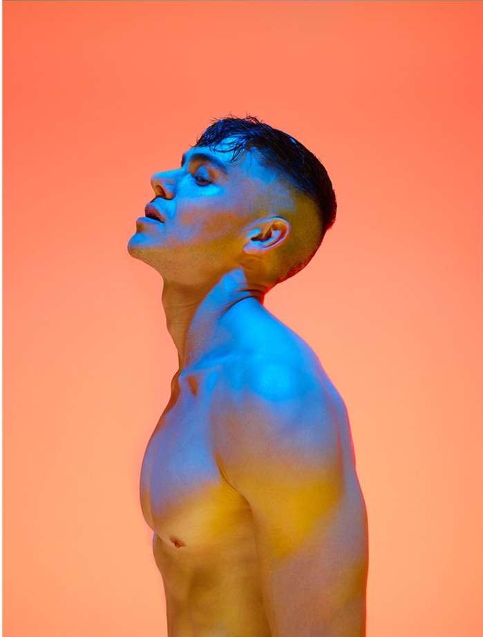 Profile portrait of a shirtless adult male with a high fade haircut, gazing upwards in a photo studio with gradient orange and blue lighting.