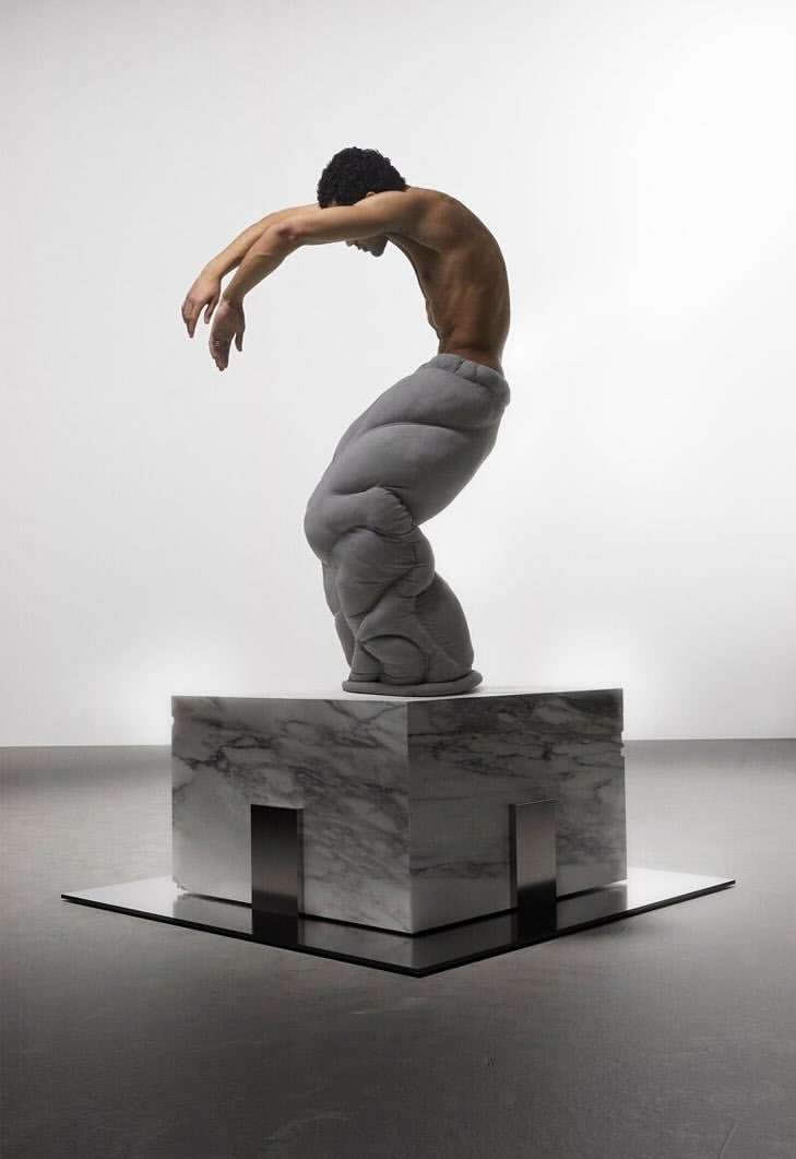 An artistic image showcasing a shirtless man in padded pants posed on a marble block, captured in a bright, minimalist studio environment.