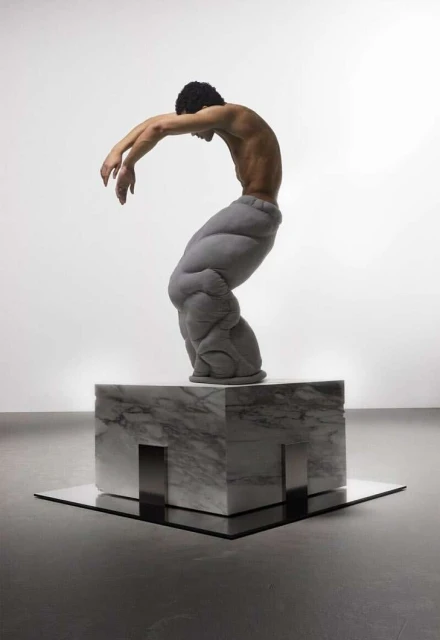 An artistic image showcasing a shirtless man in padded pants posed on a marble block, captured in a bright, minimalist studio environment.