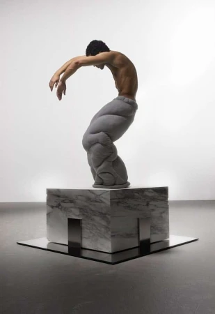 An artistic image showcasing a shirtless man in padded pants posed on a marble block, captured in a bright, minimalist studio environment.