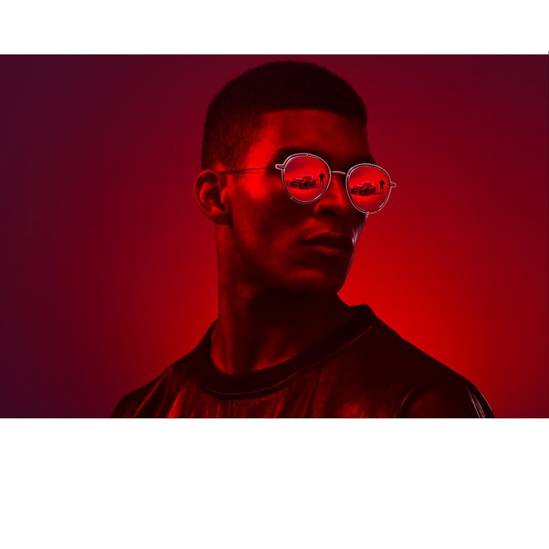 Portrait of a young adult male wearing round glasses, lit with red lighting against a dark red background, creating a striking contrast.