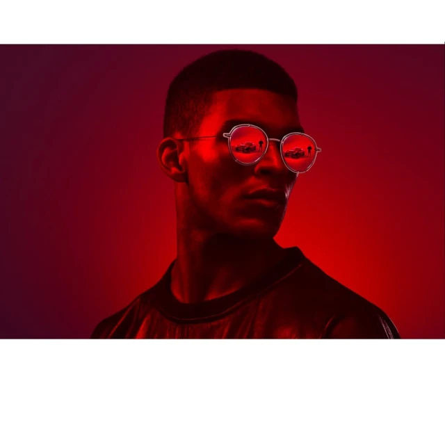 Portrait of a young adult male wearing round glasses, lit with red lighting against a dark red background, creating a striking contrast.