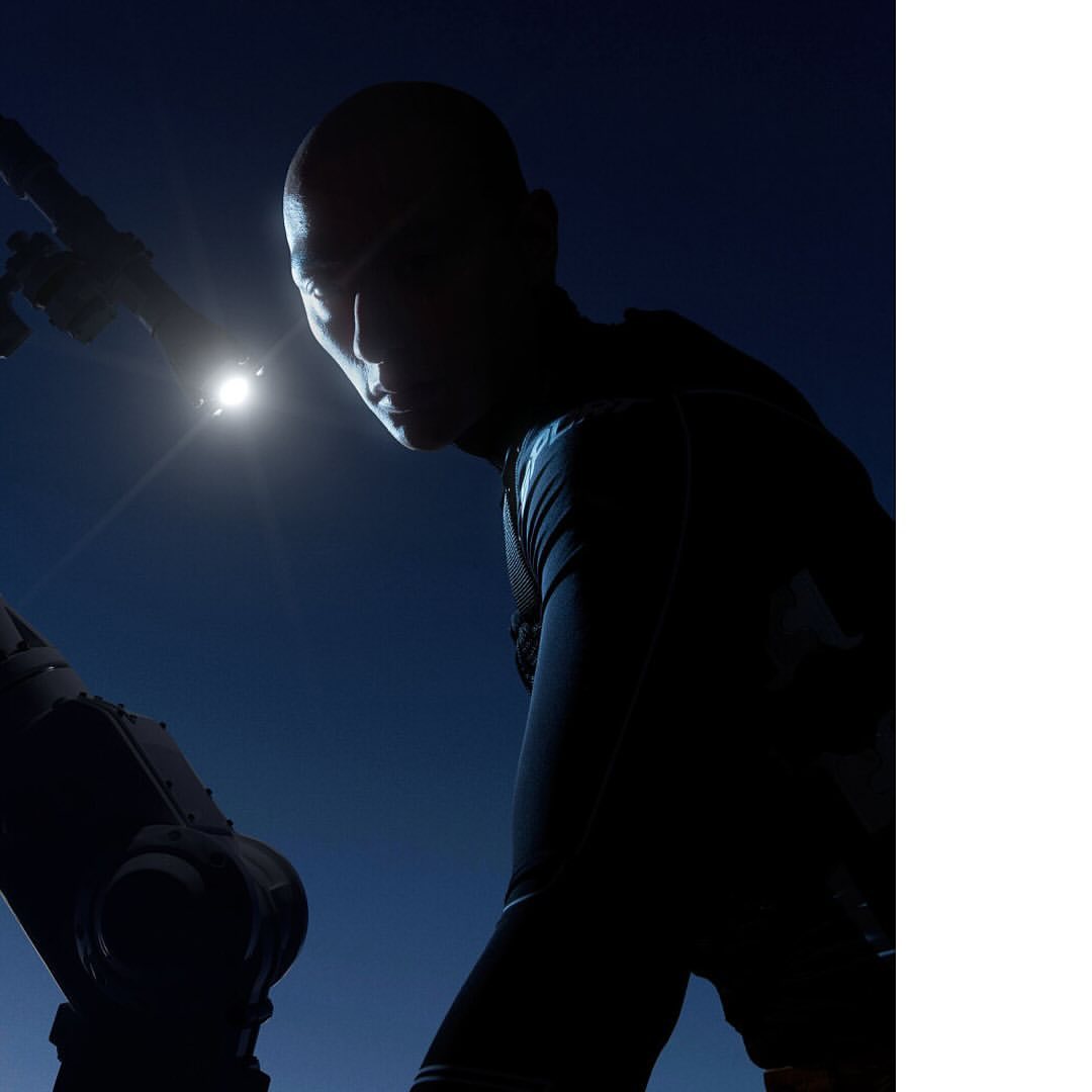 Dramatic low-angle silhouette of a bald adult male with a dark outfit under a spotlight and against a deep blue background.