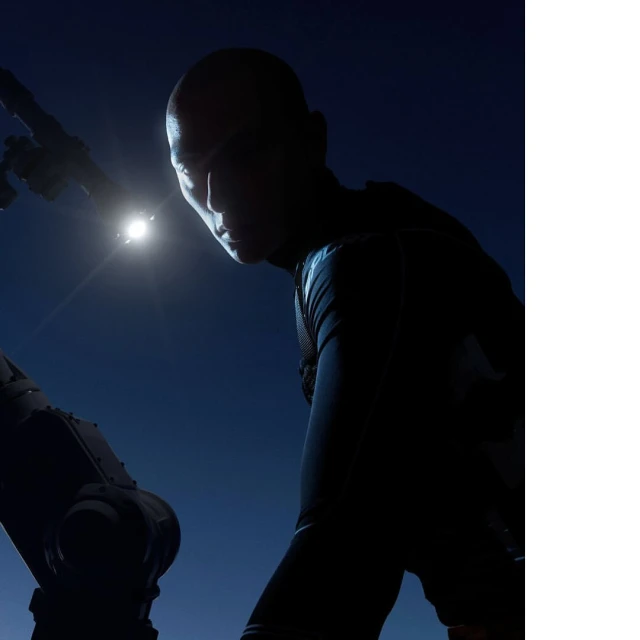 Dramatic low-angle silhouette of a bald adult male with a dark outfit under a spotlight and against a deep blue background.