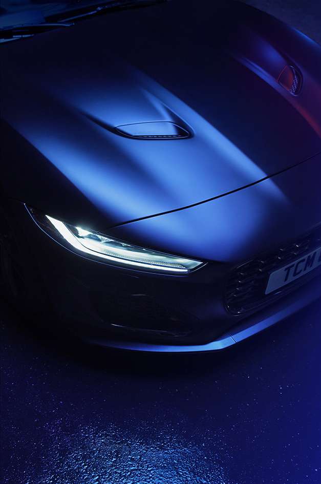 Close-up of a matte blue sports car with a visible headlight and hood vent detail under blue-purple lighting, highlighting its sleek design.