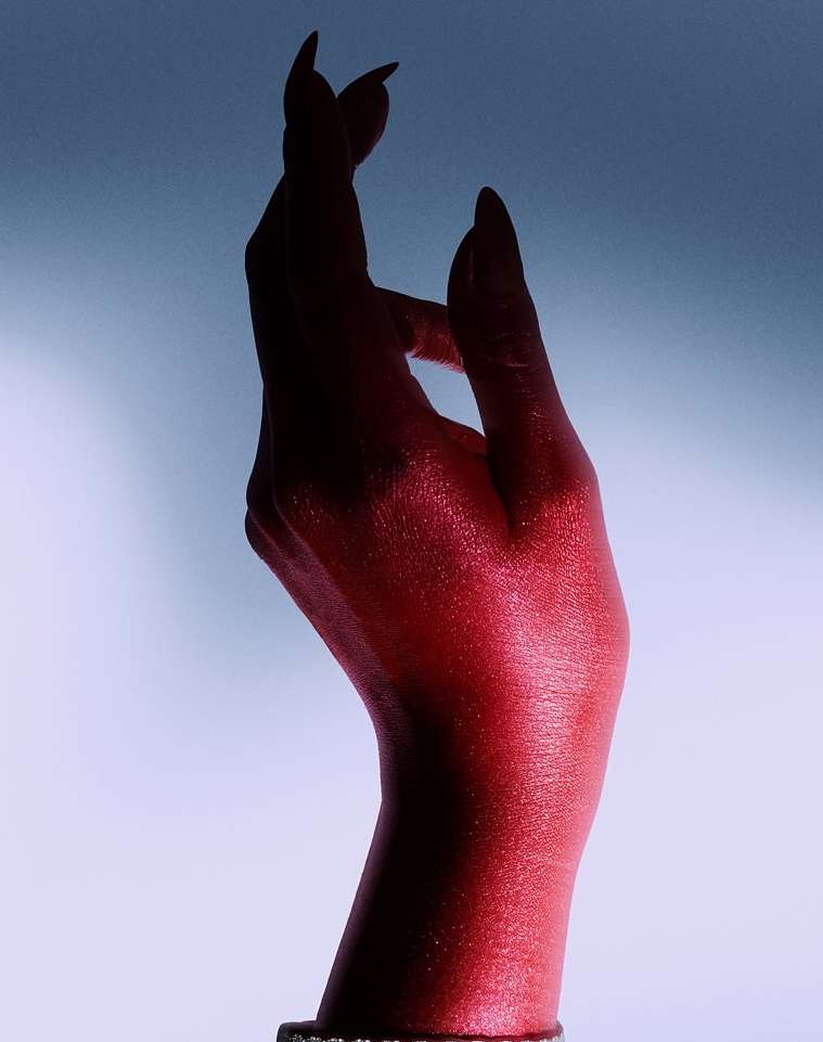 Close-up studio shot of a red hand with long painted nails against a gradient blue background. The hand is adorned with a gold bracelet.