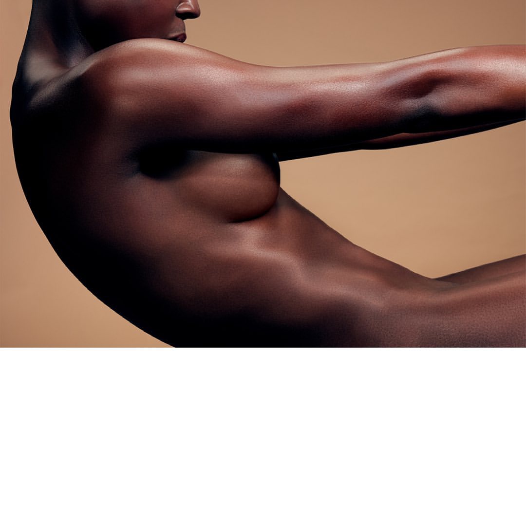 Striking studio shot of a nude adult female with dark skin, posing against a tan backdrop, showcasing her body's curves and contours.