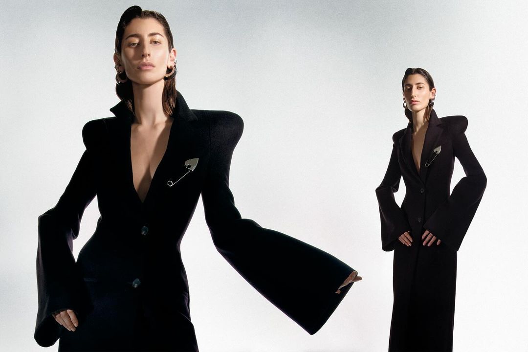 Two adult females in black formalwear with exaggerated shoulders and lapels pose against a neutral background in a fashion editorial.