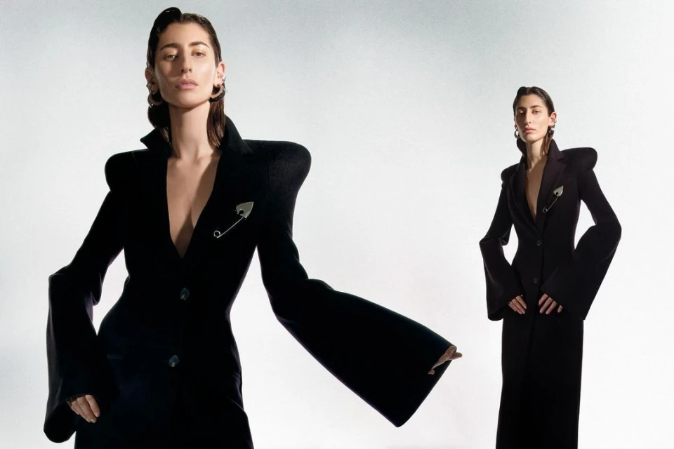 Two adult females in black formalwear with exaggerated shoulders and lapels pose against a neutral background in a fashion editorial.