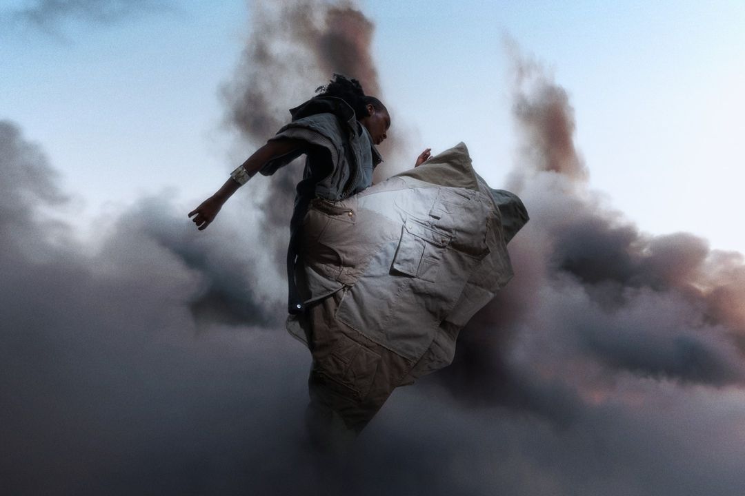 Person in stylized gray clothing stands surrounded by dark swirling smoke against a pale blue sky in a dynamic outdoor fashion shot.