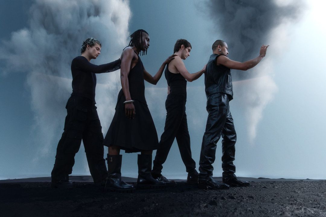 Four men in black clothing walk in a line across a black landscape. They are in a row, each with a hand on the shoulder of the person in front.