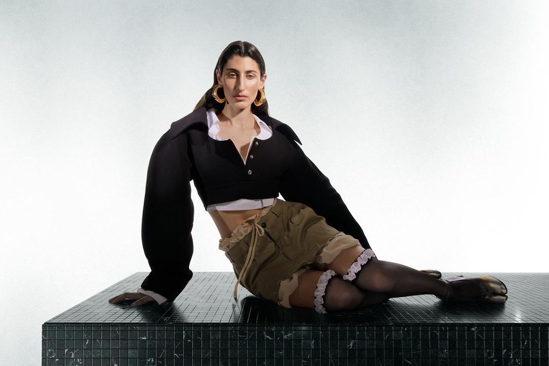 Woman in black crop top and khaki shorts sitting on a black marble surface in a studio setting with soft, diffused lighting.