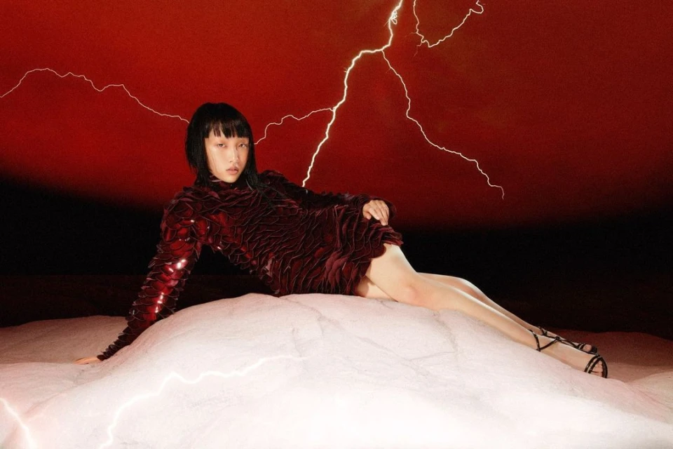 Fashionable Asian woman in a red dress reclines on a cloud-like surface against a red background with lightning details for a striking editorial.