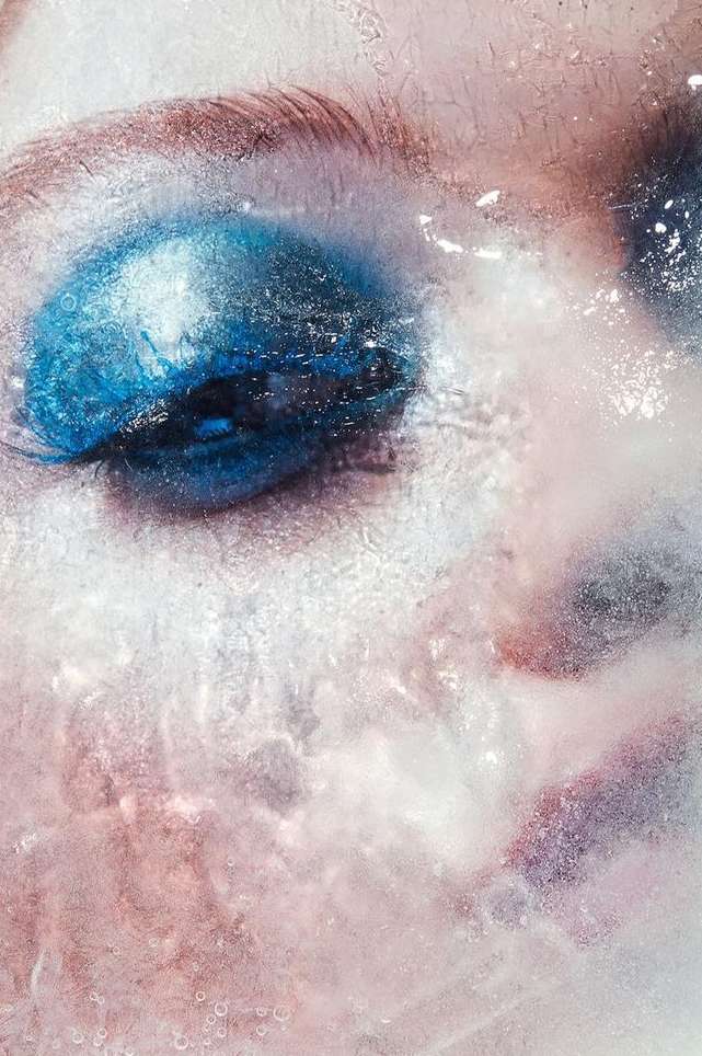 Close-up portrait of a person's face, partially covered in what appears to be water or ice, with bright blue eye makeup.