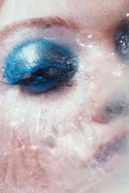Close-up portrait of a person's face, partially covered in what appears to be water or ice, with bright blue eye makeup.