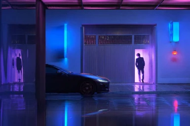 A dark sports car parked on a wet street next to a building illuminated by blue and purple neon lights with a person entering a doorway.