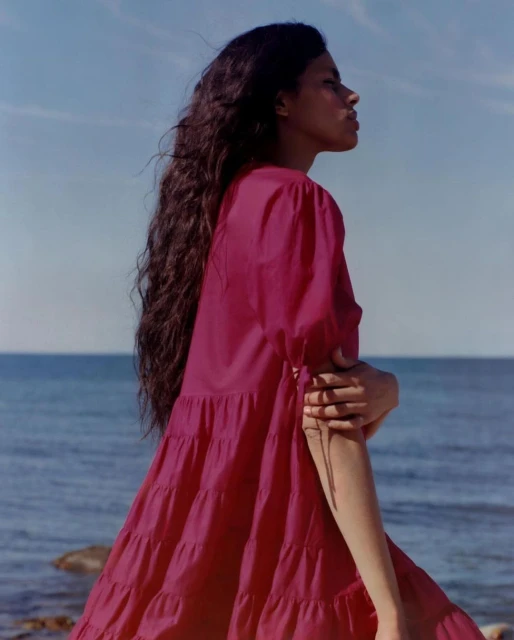 Portrait of a woman with long brown hair in a magenta dress, standing on a beach with blue water and sky in the background.