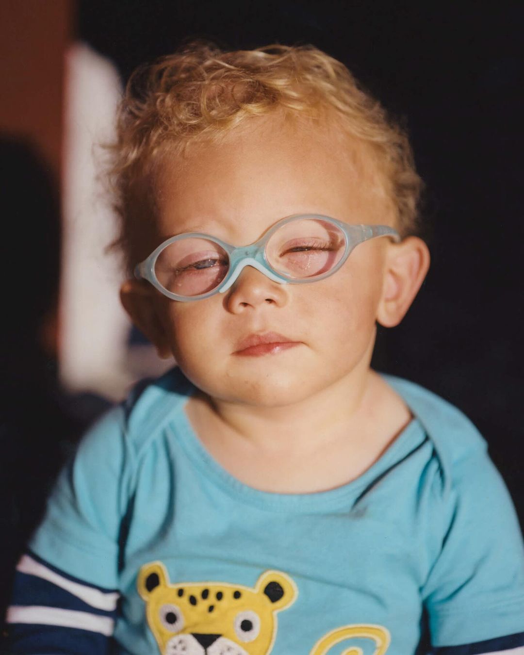 Portrait of a toddler wearing light blue glasses and a blue t-shirt with a yellow tiger on it against a dark background.