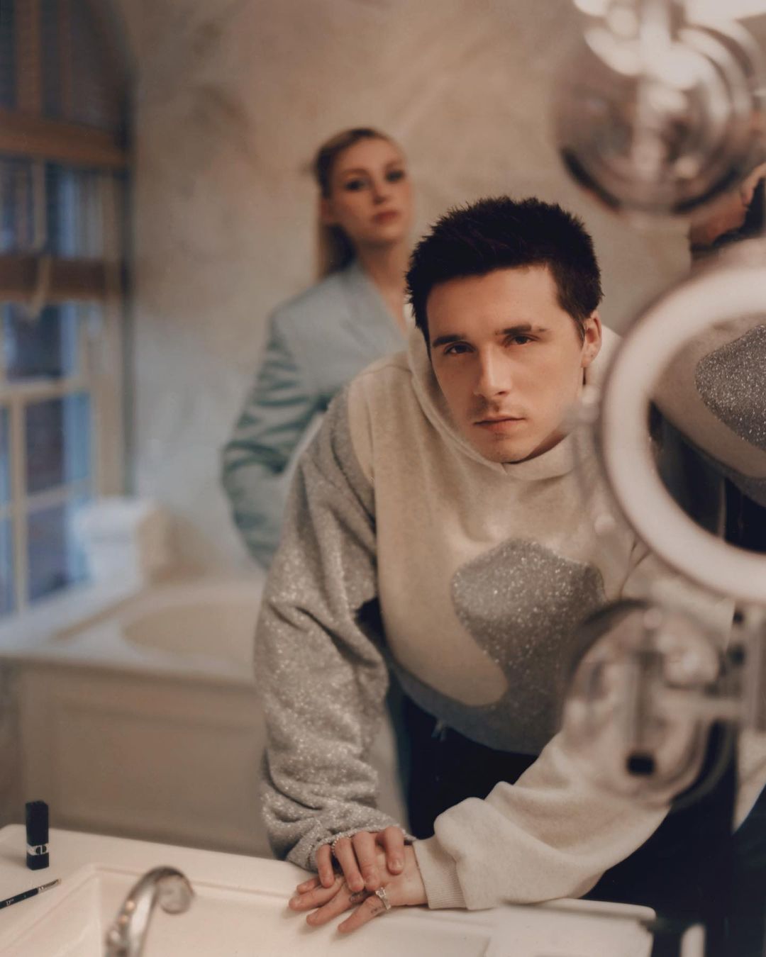 Portrait of a young adult man in a hoodie standing in a bathroom with ring light reflection, with a blurred woman in the background.