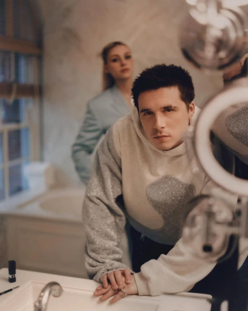 Portrait of a young adult man in a hoodie standing in a bathroom with ring light reflection, with a blurred woman in the background.