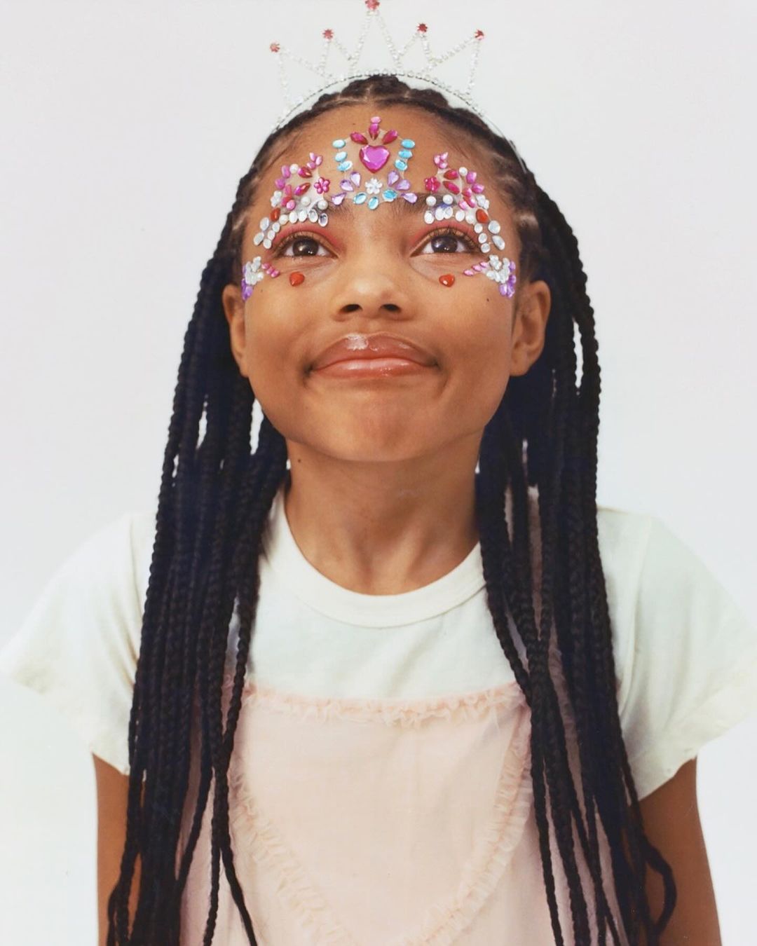 Portrait of a young girl with braided hair wearing a tiara and decorative gems on her face, looking up with a neutral expression.