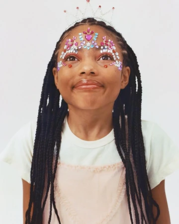 Portrait of a young girl with braided hair wearing a tiara and decorative gems on her face, looking up with a neutral expression.