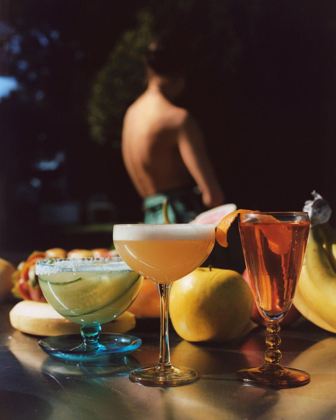 Three cocktails stand on a reflective surface with fruits, with a person blurred in background, under natural light.