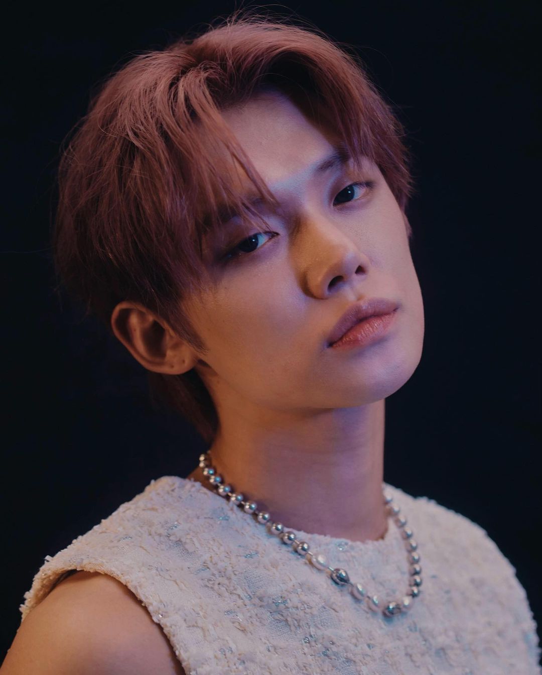 Portrait of young man with pink hair and beaded silver necklace wearing white textured sleeveless top against dark background
