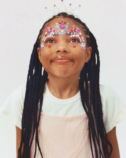 A young girl with braided hair smiles at the camera. She has decorative face jewels and a tiara in front of a white backdrop.