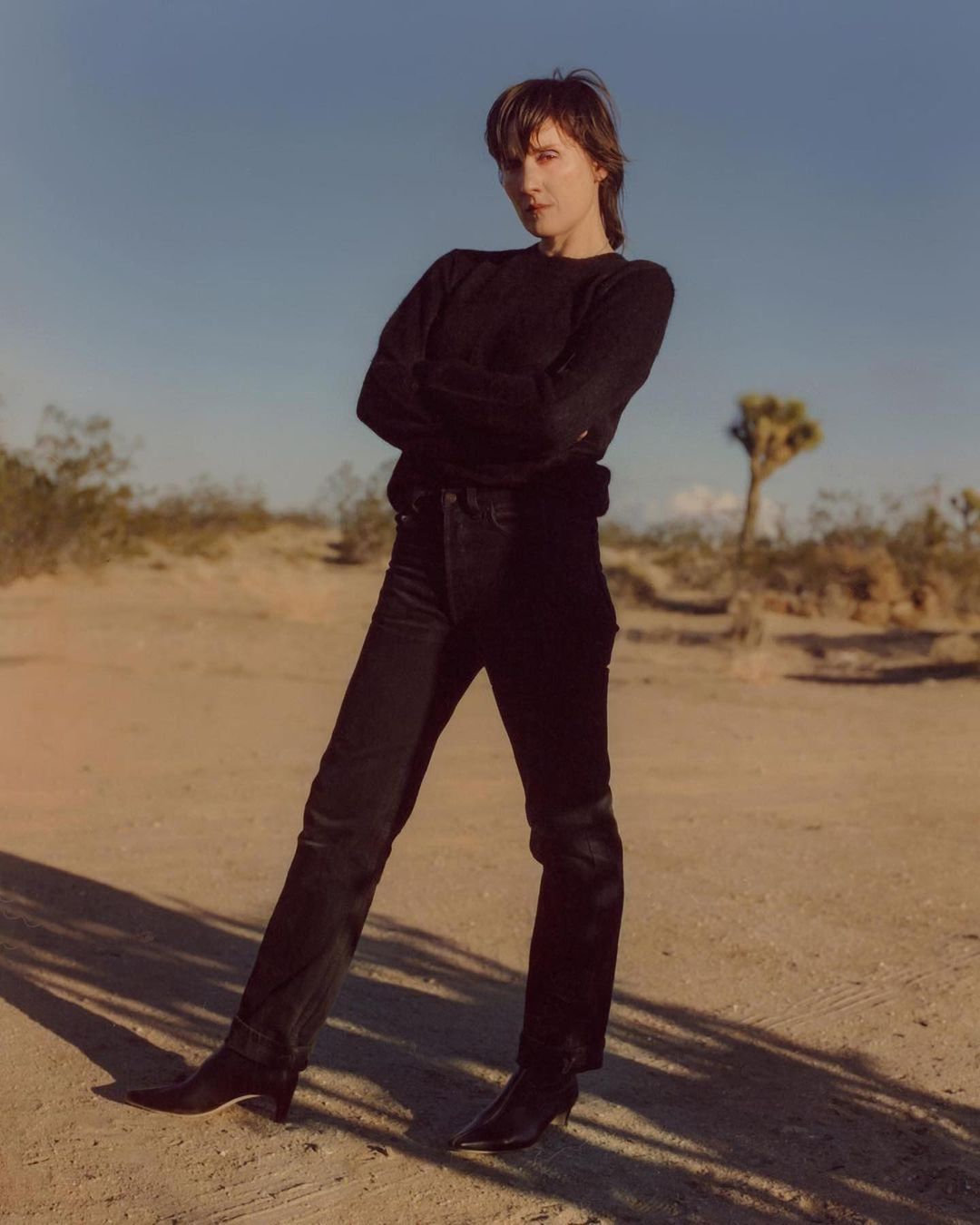 Woman poses in black sweater and jeans in the desert with sparse vegetation under a clear sky, standing in front of desert landscape.