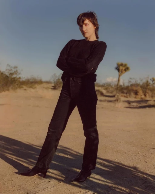 Woman poses in black sweater and jeans in the desert with sparse vegetation under a clear sky, standing in front of desert landscape.