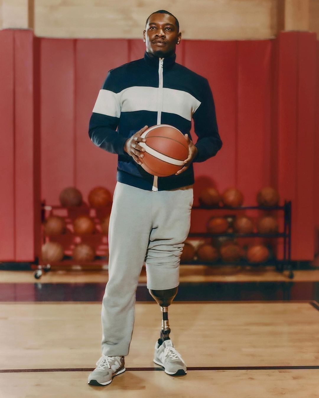 A man with a prosthetic leg standing indoors and holding a basketball. He is in athletic wear with a wooden court behind him.