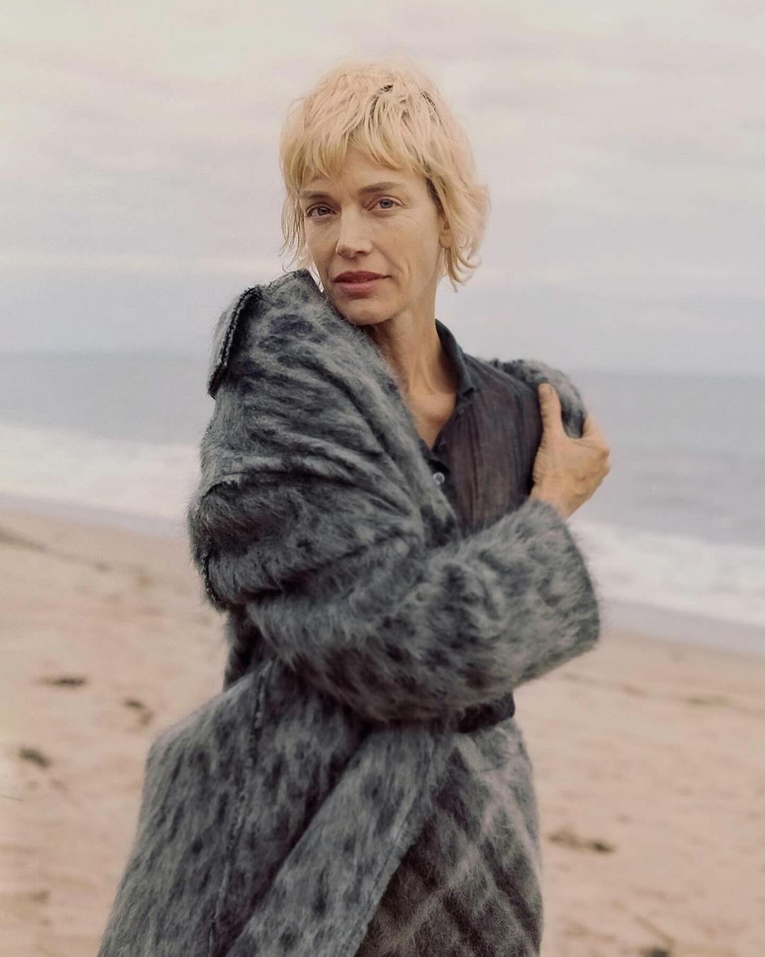 Adult woman posing on a sandy beach wearing an oversized grey coat and a dark top, with the ocean and cloudy sky in the background.
