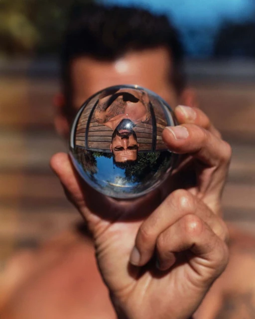 Adult male holds a glass sphere reflecting the sky and trees, with an out-of-focus face visible in the background.