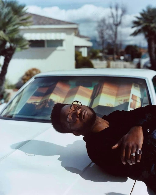 Adult man with glasses, wearing a dark shirt and rings, reclines on the hood of a white car on a sunny day in a residential neighborhood.