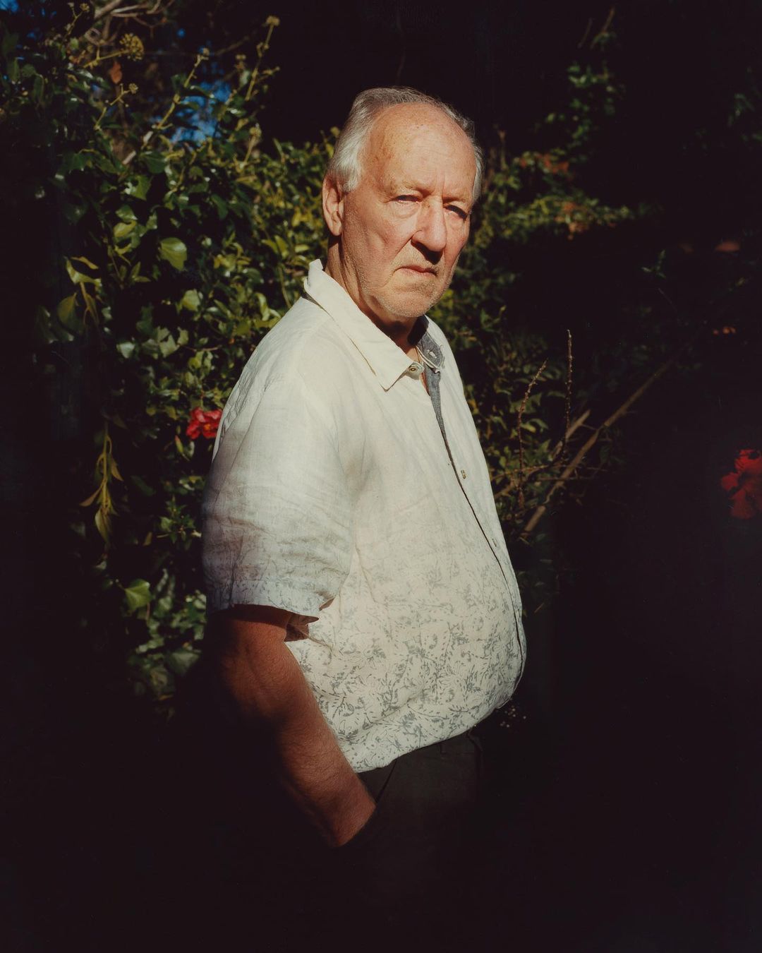 Portrait of a mature adult man outdoors, wearing a patterned white short sleeve shirt, standing in front of a lush green bush with red flowers.