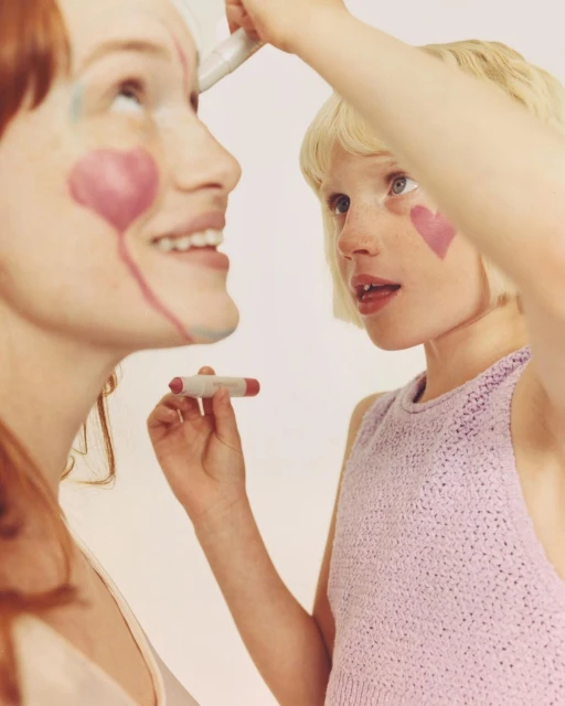 Young girl applies heart-shaped face paint on adult woman in a close up studio portrait with soft lighting against a white background.