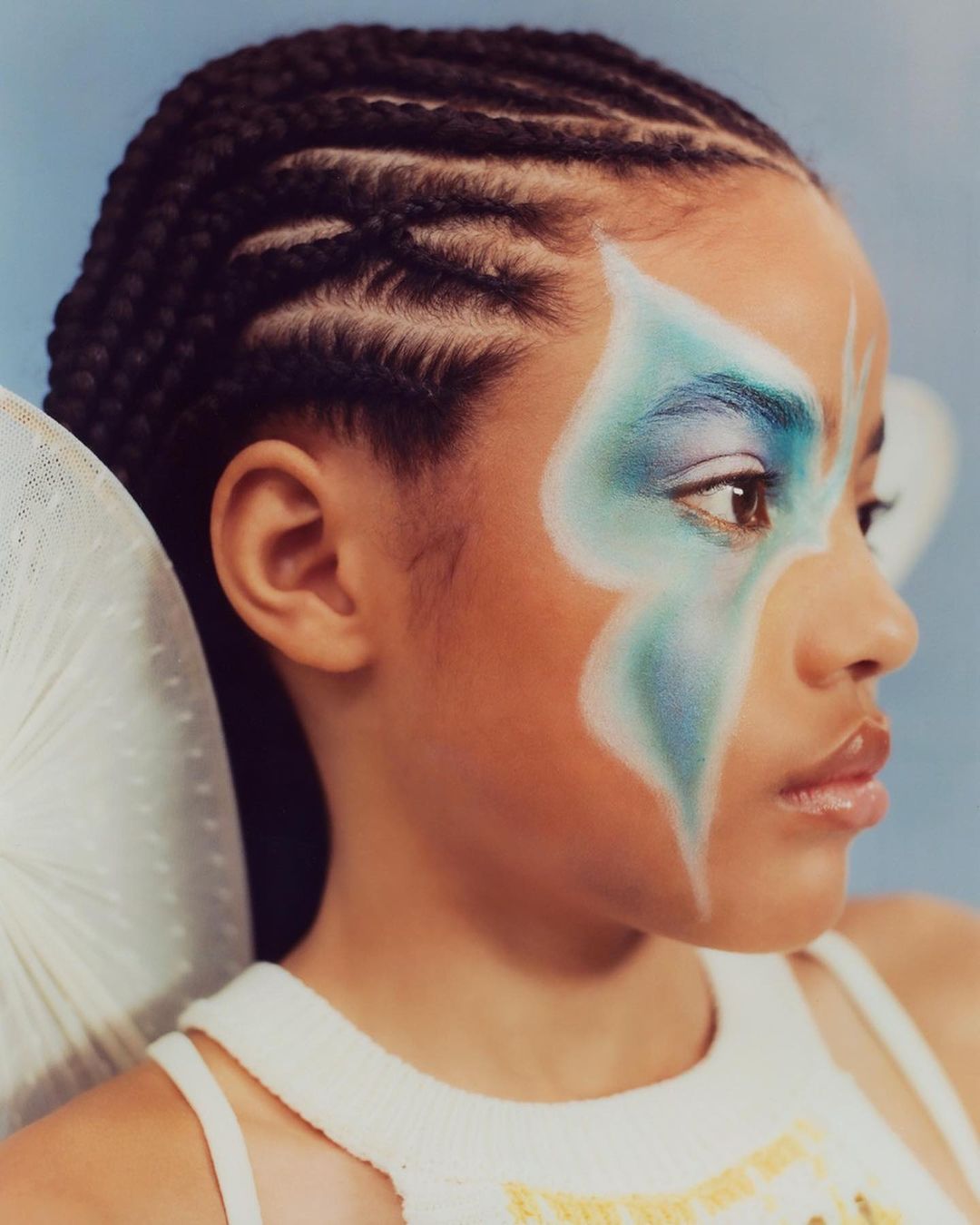 Close-up portrait of a girl with braided hair and blue face paint, showcasing beauty makeup against a soft blue background.