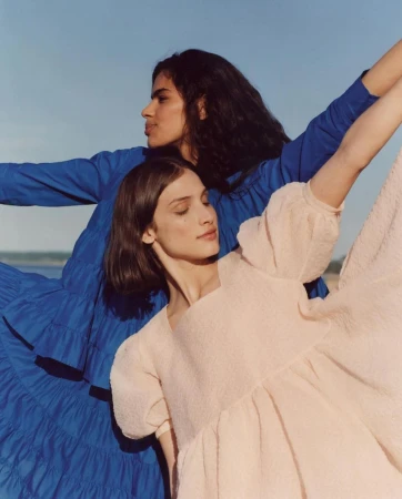 Two women posing outdoors against a clear sky, one wearing a blue dress and the other in a light pink dress