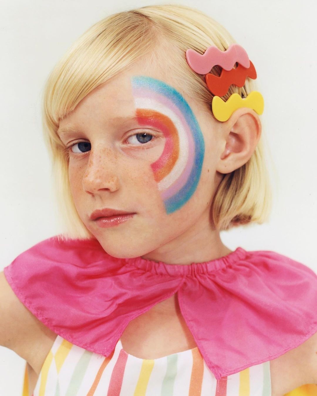 Close-up portrait of a child with blonde hair, wearing colorful face paint and hair clips, posing against a neutral background.