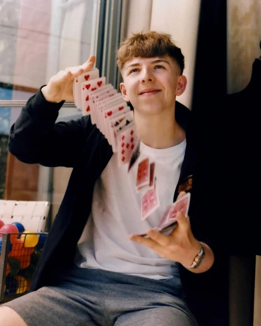Young man sits indoors juggling playing cards in the air, wearing a black jacket, gray shorts, and white shirt. Window in background.