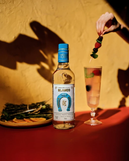 Bottle of Herradura Blanco Tequila next to a cocktail with berries, garnished with a skewer of fresh blackberries and strawberries.