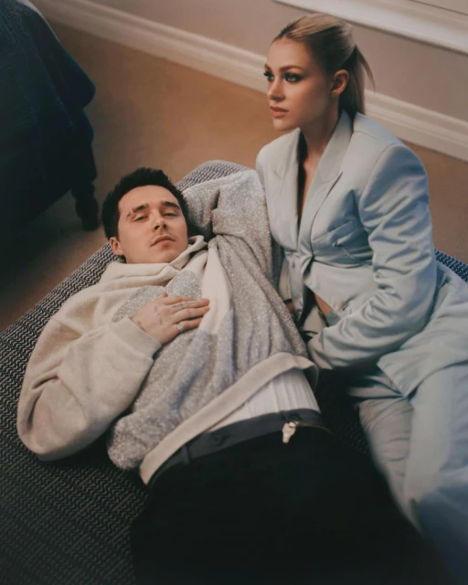 A couple relaxes indoors; the man lies down while the woman in a blazer sits beside him in a softly lit home environment.