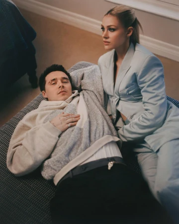 A couple relaxes indoors; the man lies down while the woman in a blazer sits beside him in a softly lit home environment.