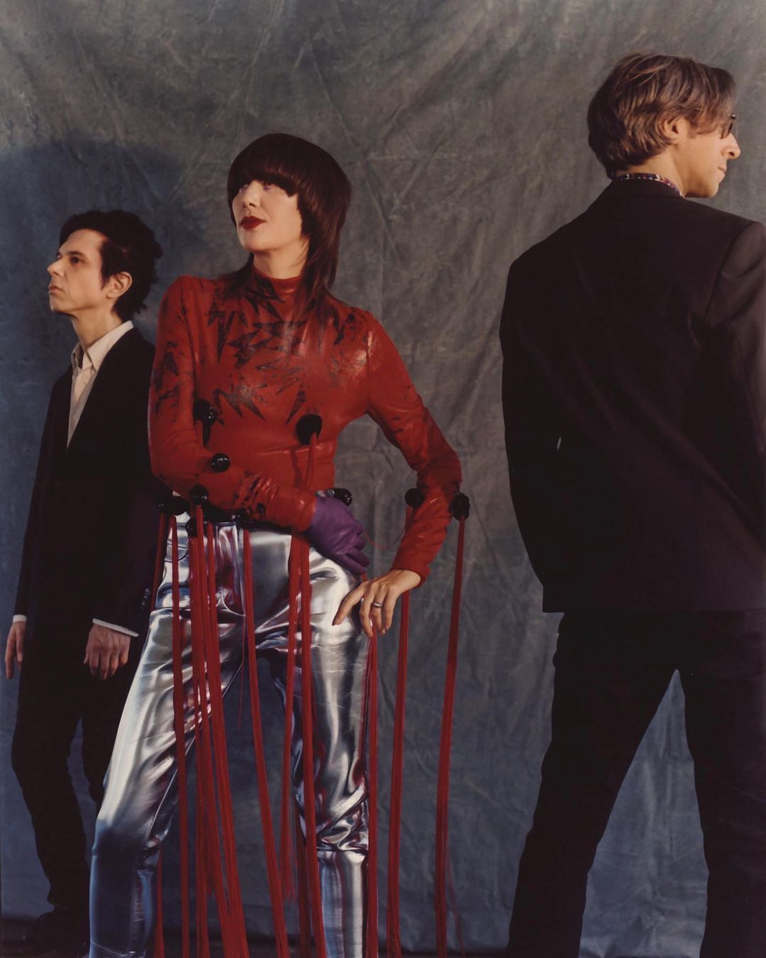 Three people pose against a grey mottled backdrop: a woman in red with silver pants and two men in black suits.