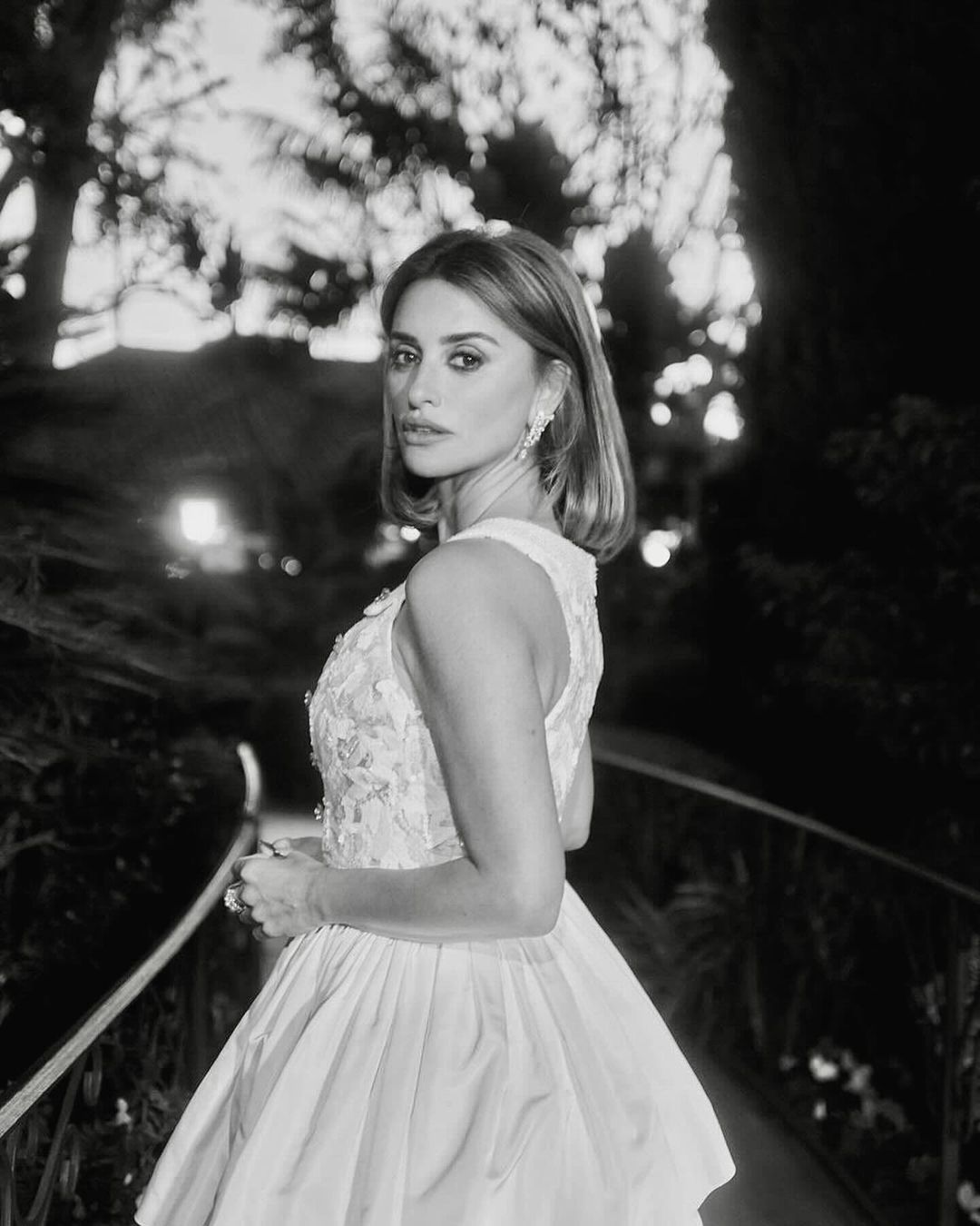 Penélope Cruz gazes over her shoulder in an eye-level medium shot, wearing an off-white gown with floral embellishments outdoors.