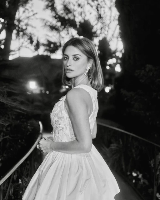 Penélope Cruz gazes over her shoulder in an eye-level medium shot, wearing an off-white gown with floral embellishments outdoors.